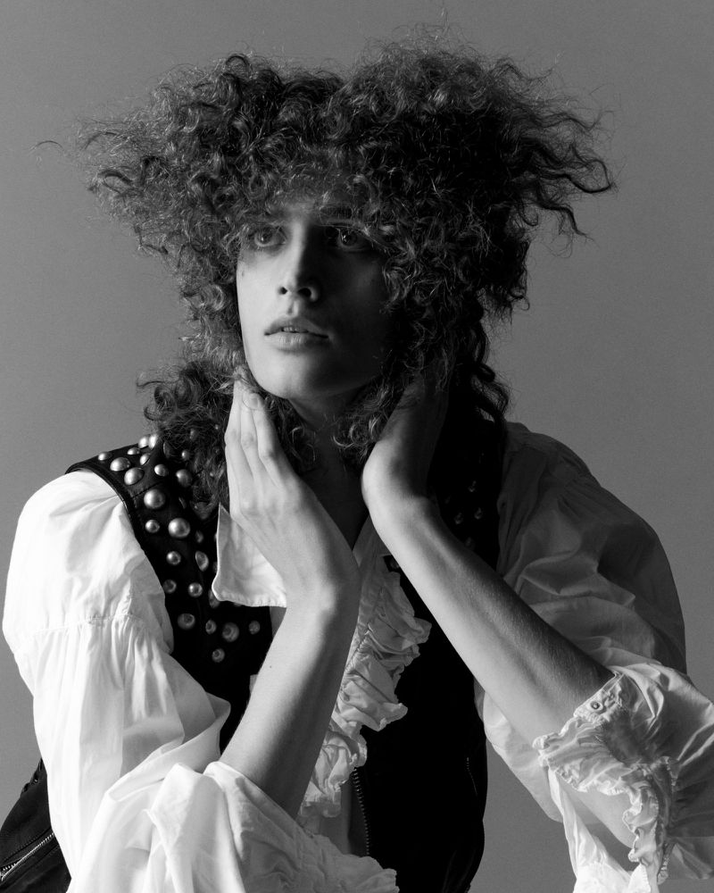 Black and white portrait of a person with curly hair wearing a studded vest over a ruffled shirt, posing with hands near the neck.