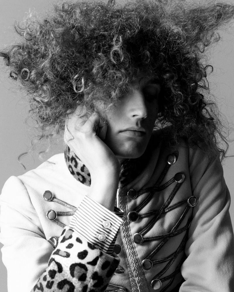 Black-and-white portrait of a person with afro, wearing a decorated jacket with animal-print accents and posing indoors.