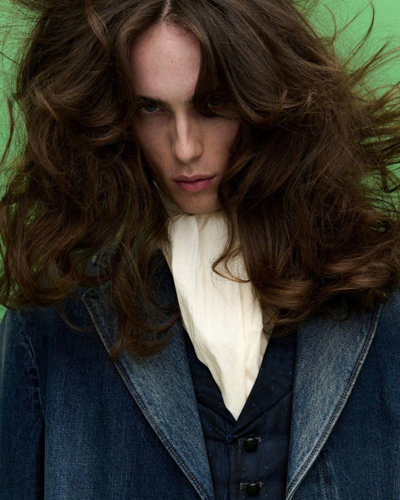 Portrait of a young adult with long brown wavy hair, wearing a denim jacket and white collared shirt, against a vibrant green backdrop.