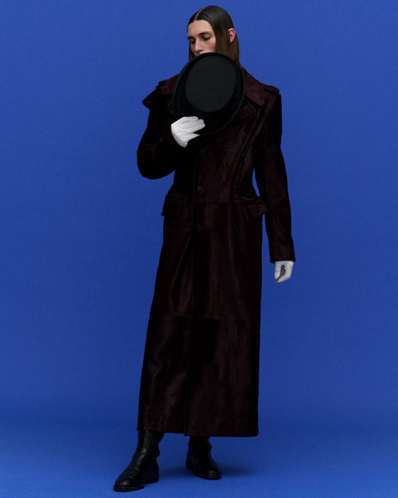 Elegant man poses in a long dark coat and gloves against a bright blue backdrop, holding a black bowler hat in a portrait-style shot.