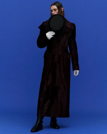 Elegant man poses in a long dark coat and gloves against a bright blue backdrop, holding a black bowler hat in a portrait-style shot.