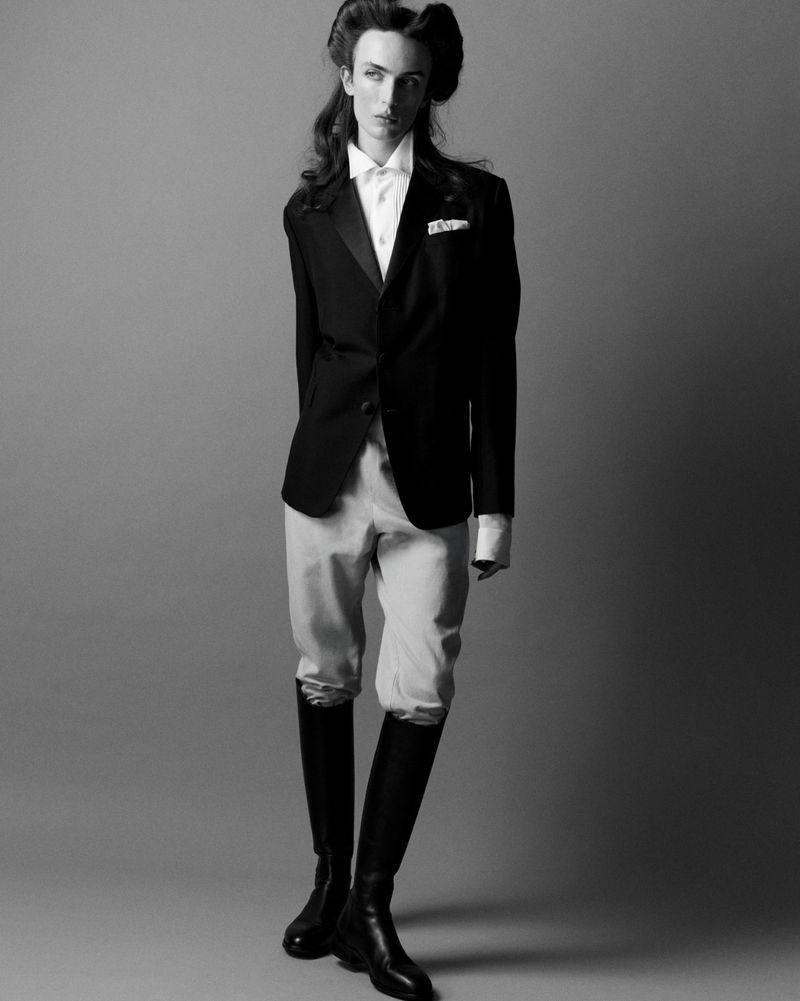 Black and white portrait of a person in a black jacket, white shirt, riding pants and boots in a minimalist studio setting.