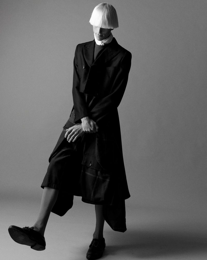 Monochrome fashion shot of a slender person in a dark overcoat, wide shorts, and blunt blonde wig, posing in a studio.