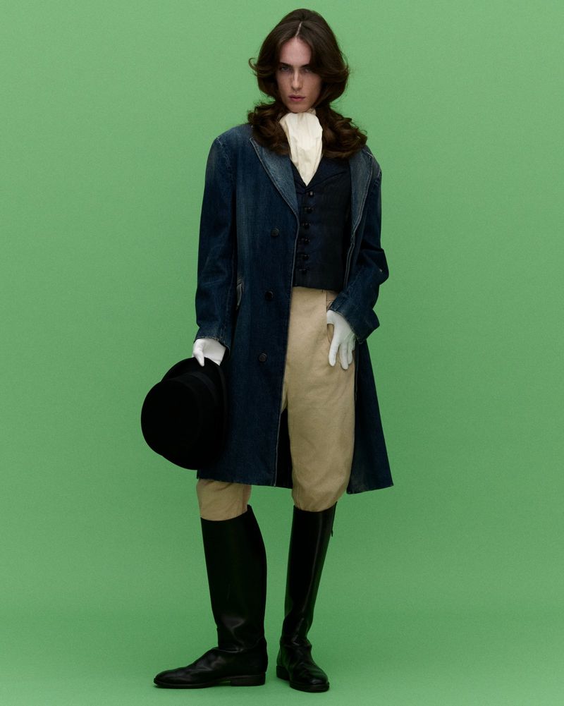 Full-length portrait of a man in a vintage-inspired outfit: long blue coat, dark vest, riding pants, and boots against a green backdrop.