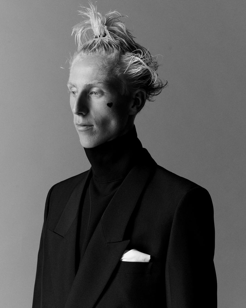 Monochrome portrait of an adult male in a black turtleneck and suit, gazing off-camera with minimalist heart tattoo on his cheek.