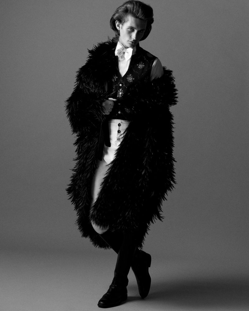 Black and white portrait of a fashionable adult male in a waistcoat, dress shirt, dark coat, black knee-high boots, and light trousers.