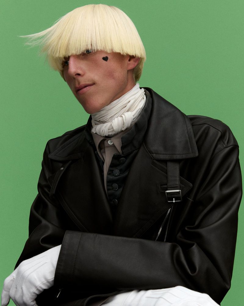 Portrait of a stylish adult male with a blonde bowl cut and a black heart tattoo wearing a black coat against a green backdrop.