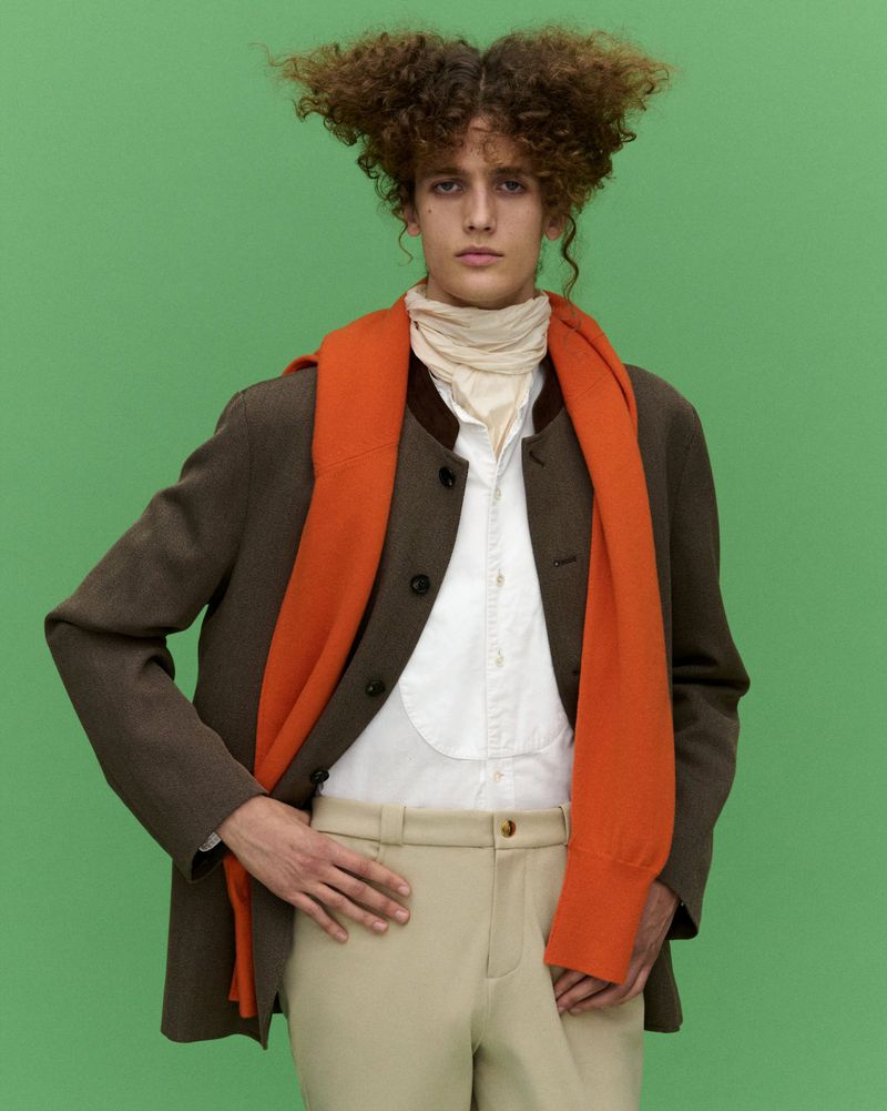 A young adult poses in tan pants, white button-down, burnt orange scarf, and brown jacket against a solid green backdrop.