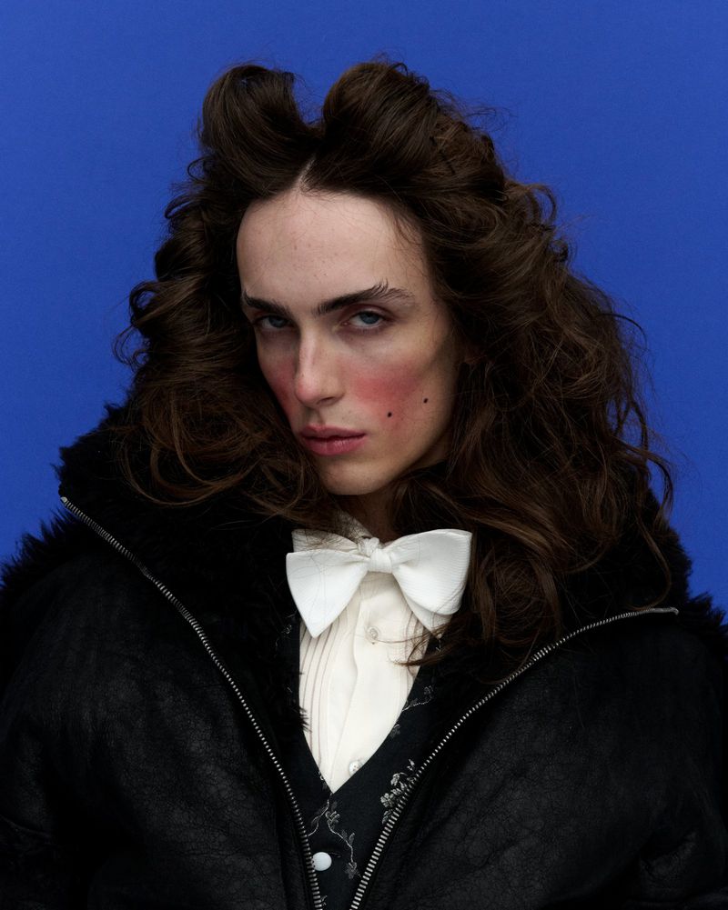 Close-up portrait of a person with long, wavy brown hair, wearing a black jacket and white bow tie against a blue backdrop.
