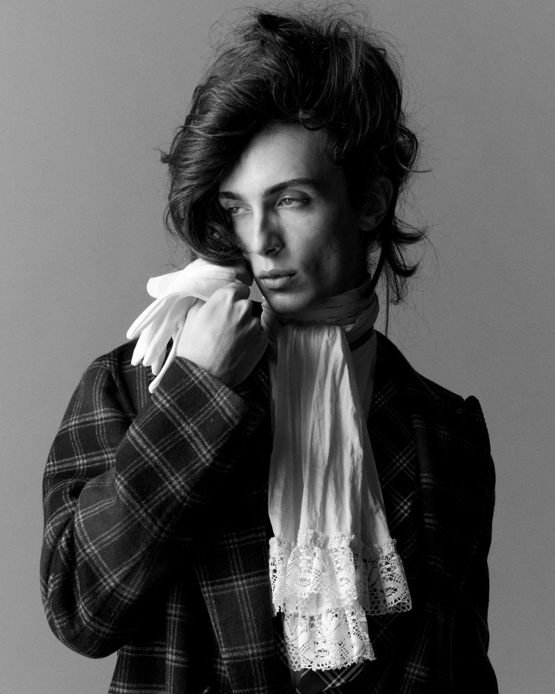 Model in plaid jacket holding white gloves with a lace cravat against grey backdrop for Arena Homme +