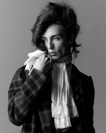 Model in plaid jacket holding white gloves with a lace cravat against grey backdrop for Arena Homme +
