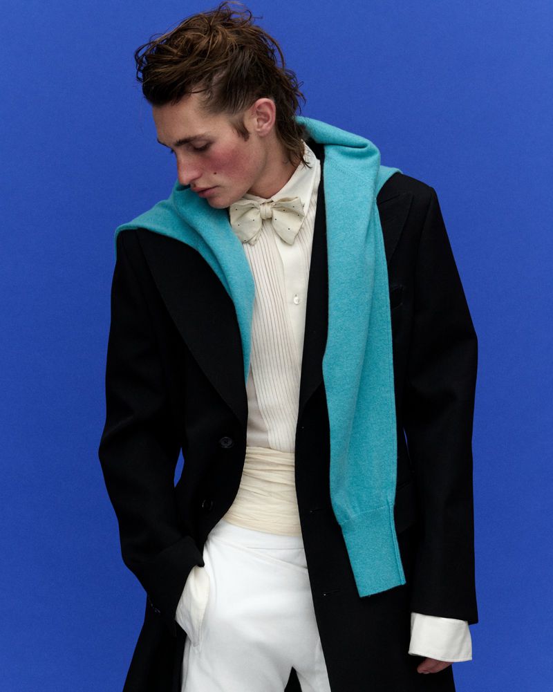 Model in black jacket with a turquoise sweater draped over shoulders and white shirt against a blue background