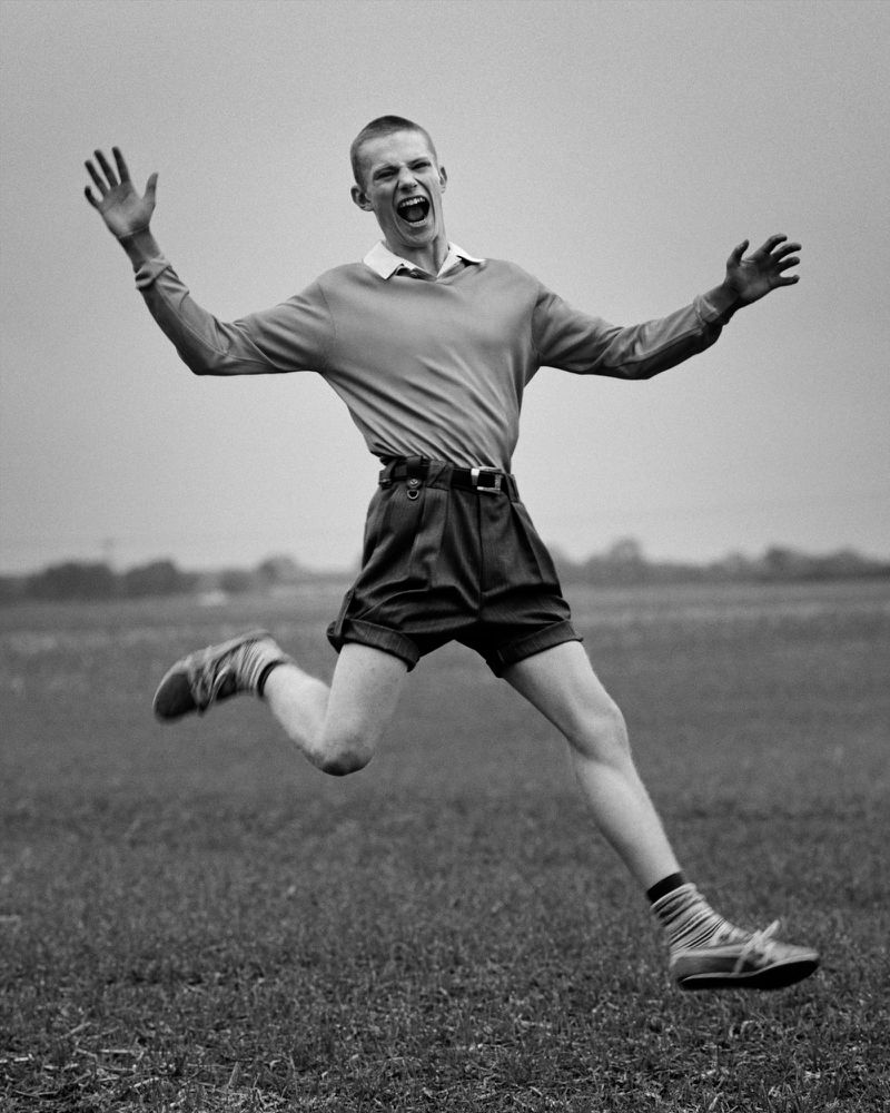 Monochrome portrait of a young man in mid-air, jumping joyfully with open arms and a wide smile in an outdoor grassy field.