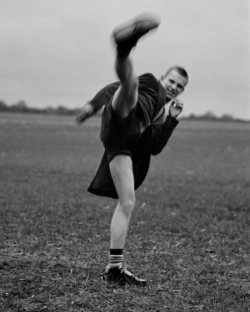 Black and white portrait of a young adult man in a suit jacket high-kicking in a grassy field on an overcast day.