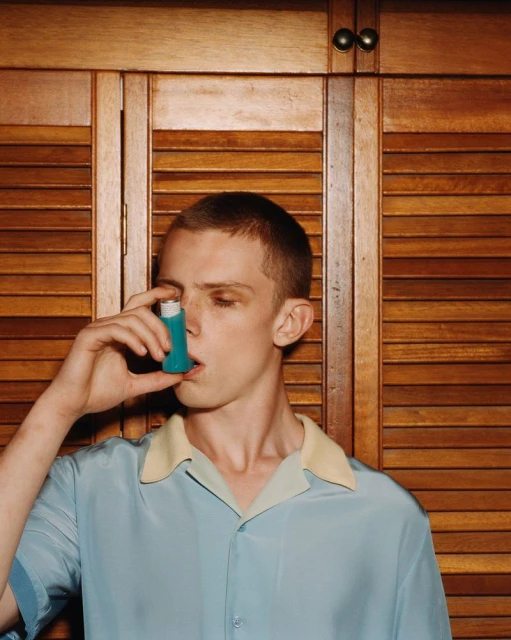 Young man uses a blue inhaler indoors, wearing a collared shirt and standing against a slatted wood background.