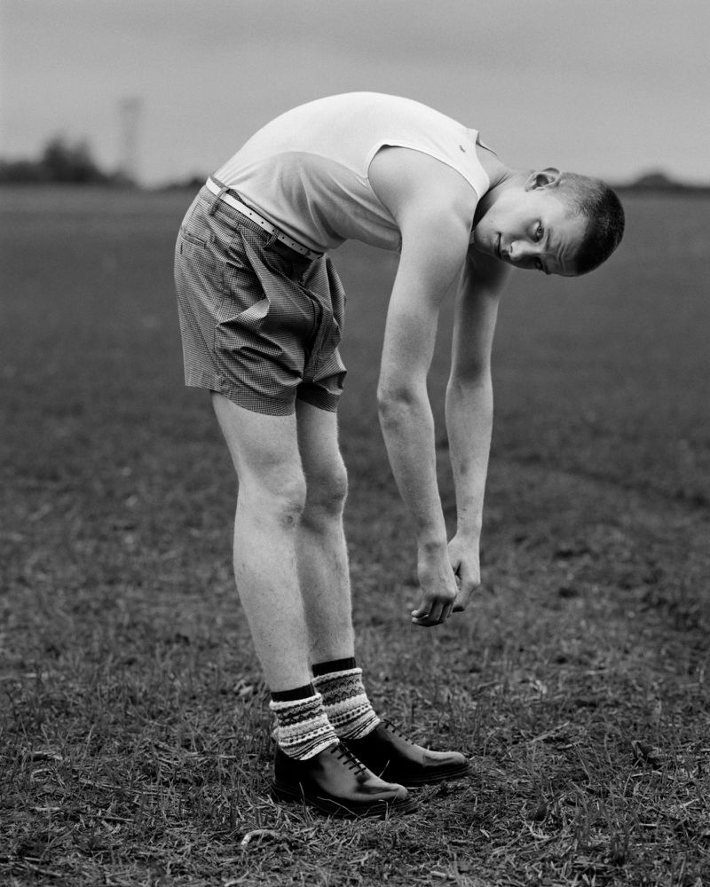 Black and white image of a male with a short haircut, wearing shorts and dress shoes, bending over backward in a grassy field.