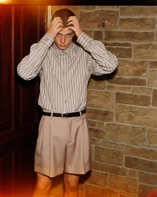 Portrait of a young adult man with hands on his head, wearing a striped shirt and khaki shorts, posing against a textured stone wall indoors.