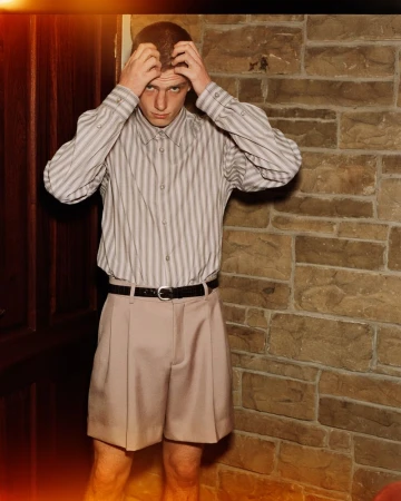 Portrait of a young adult man with hands on his head, wearing a striped shirt and khaki shorts, posing against a textured stone wall indoors.