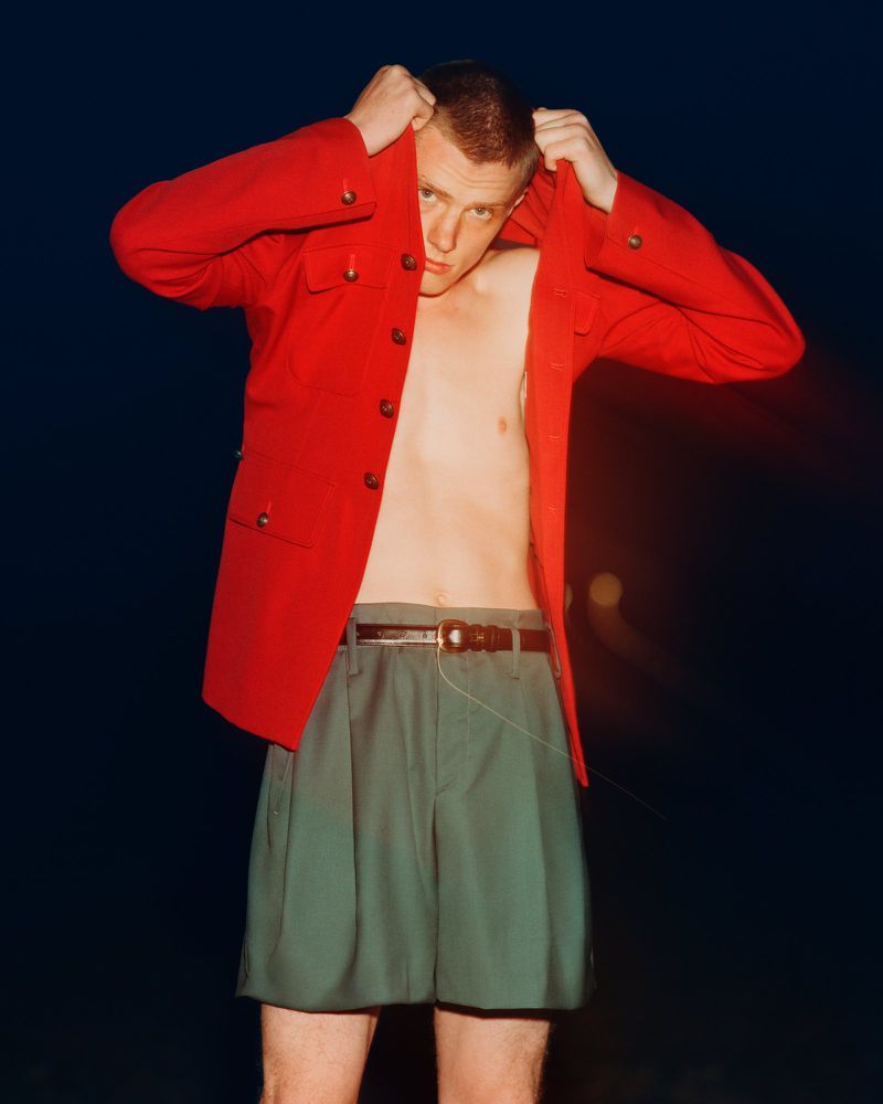 Young man poses in red jacket and green shorts in a fashion shot on a dark background with soft red lighting accents. 