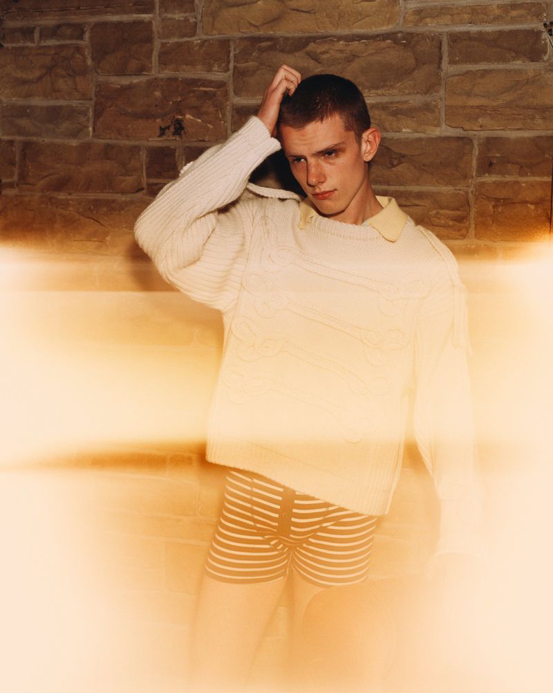 Young adult male in sweater and striped shorts poses in front of a stone wall with golden light flares adding a vintage feel.