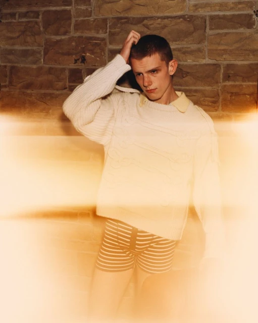 Young adult male in sweater and striped shorts poses in front of a stone wall with golden light flares adding a vintage feel.