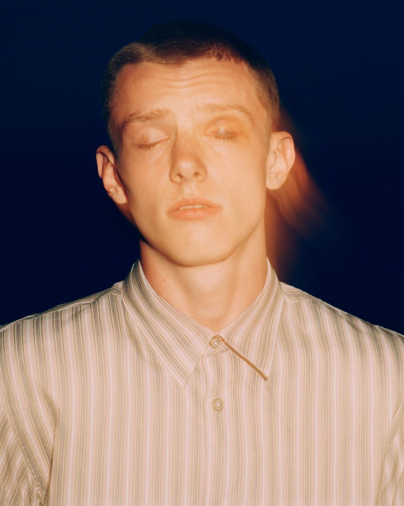 Close-up portrait of a young adult male with closed eyes, short hair, and wearing a striped button-down shirt against a dark backdrop.