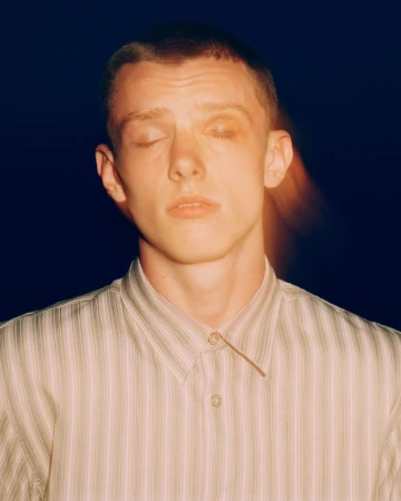 Close-up portrait of a young adult male with closed eyes, short hair, and wearing a striped button-down shirt against a dark backdrop.