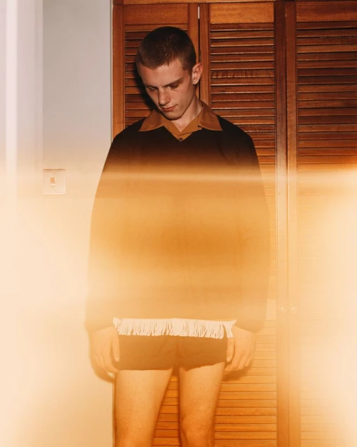 Young adult male in a brown and black sweater and black shorts poses indoors in front of a wooden door in a medium shot.