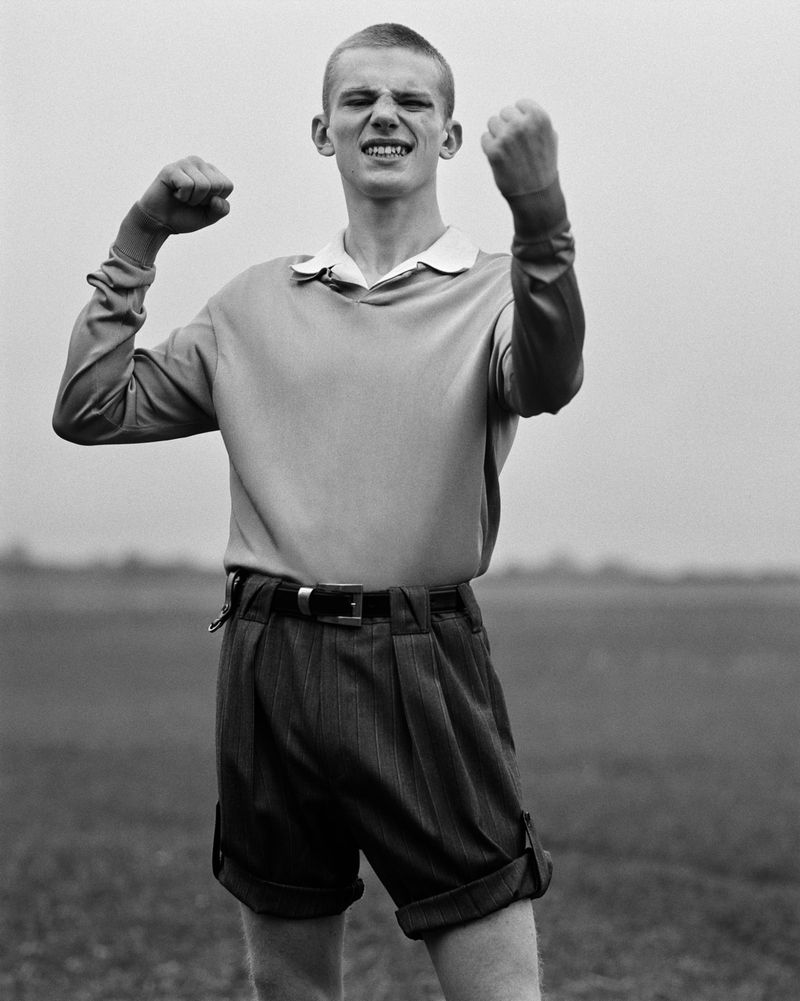 Young man in vintage-style shorts and sweater stands outdoors with fists clenched, set against a neutral grassy background and overcast sky.