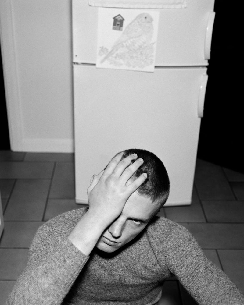 Monochrome portrait of a young adult man with short hair, wearing a sweater, seated indoors with hand on his forehead. 