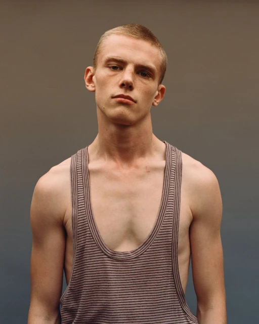 Portrait of a young adult male with short hair, wearing a striped tank top, against a blurred studio background.