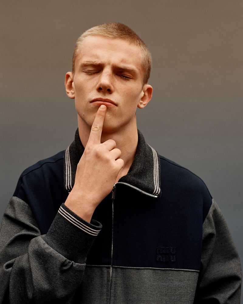 Close-up of a young adult man with a buzzcut and his eyes closed, wearing a two-tone track jacket with a finger touching his lips.