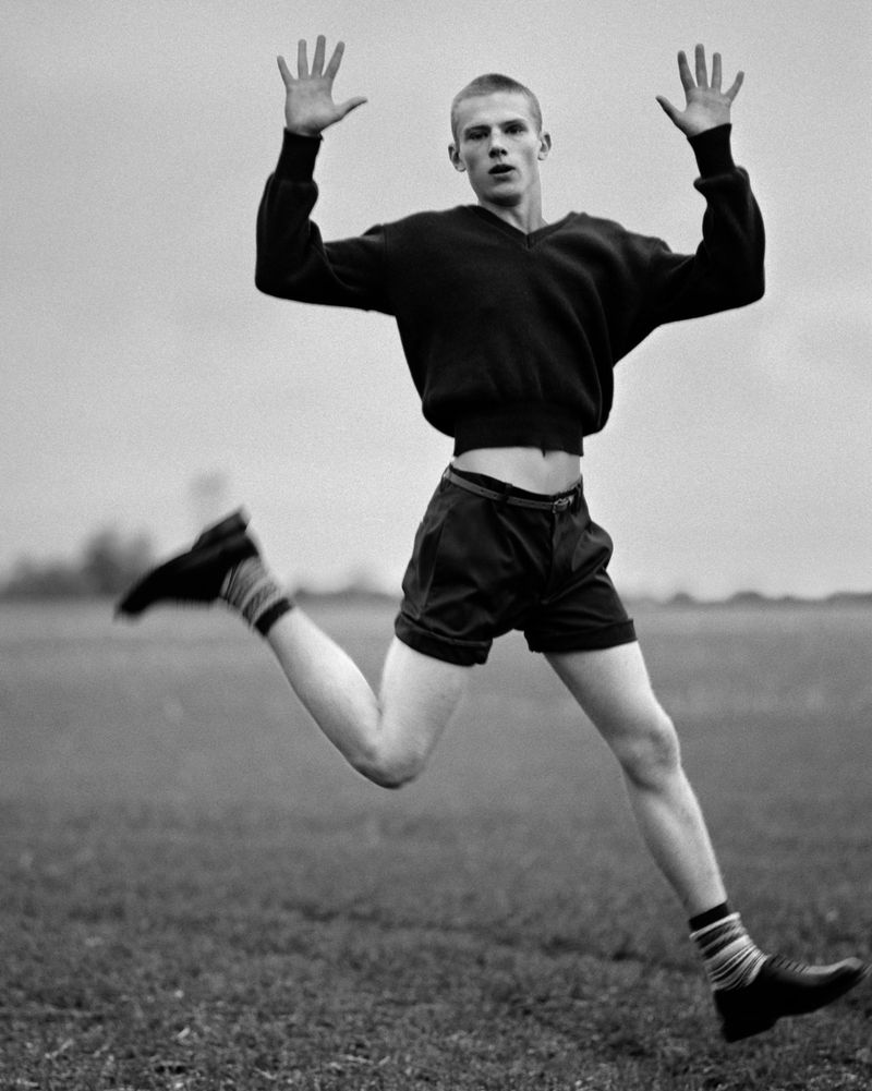 Black and white portrait of a young man jumping in the air with his hands raised, wearing a sweater, shorts, striped socks and shoes.