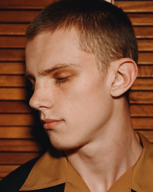 Close-up portrait of a young adult male with short brown hair, tan skin, and brown eyes, posed against a wood-paneled background indoors.