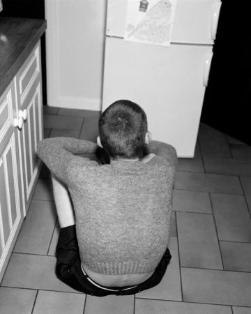 A person sits on a tiled kitchen floor with their arms crossed over their knees, facing a white refrigerator in a black and white shot.