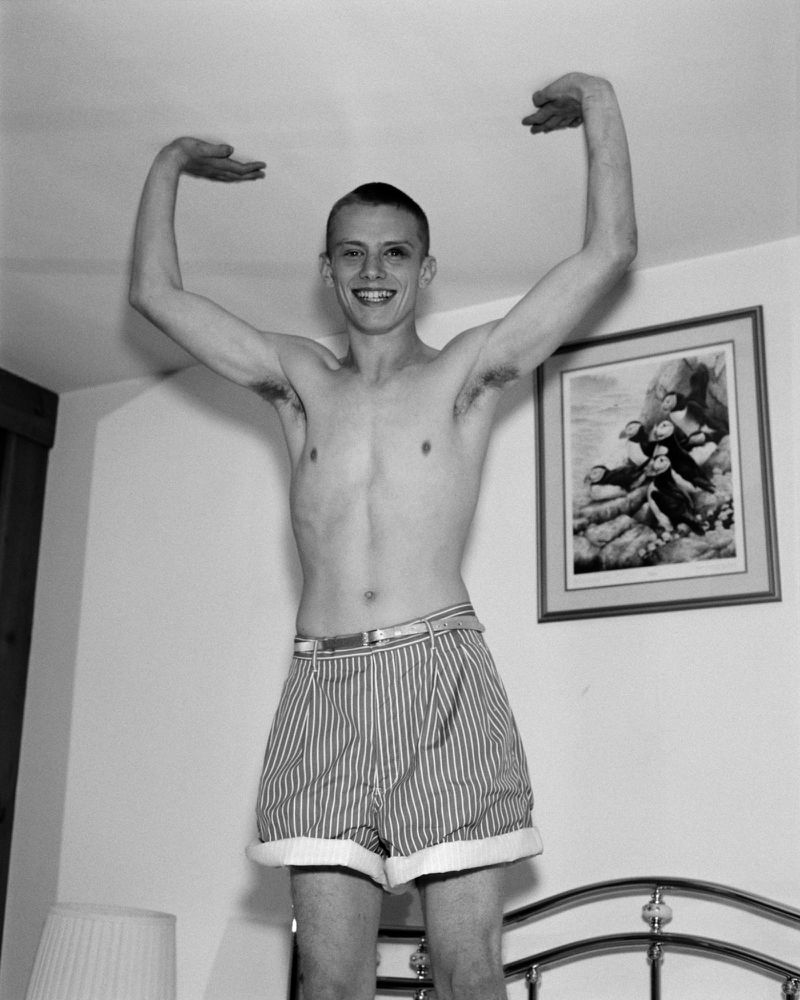 Black and white portrait of shirtless man in striped shorts striking a pose in an indoor setting with a bed and picture frame.