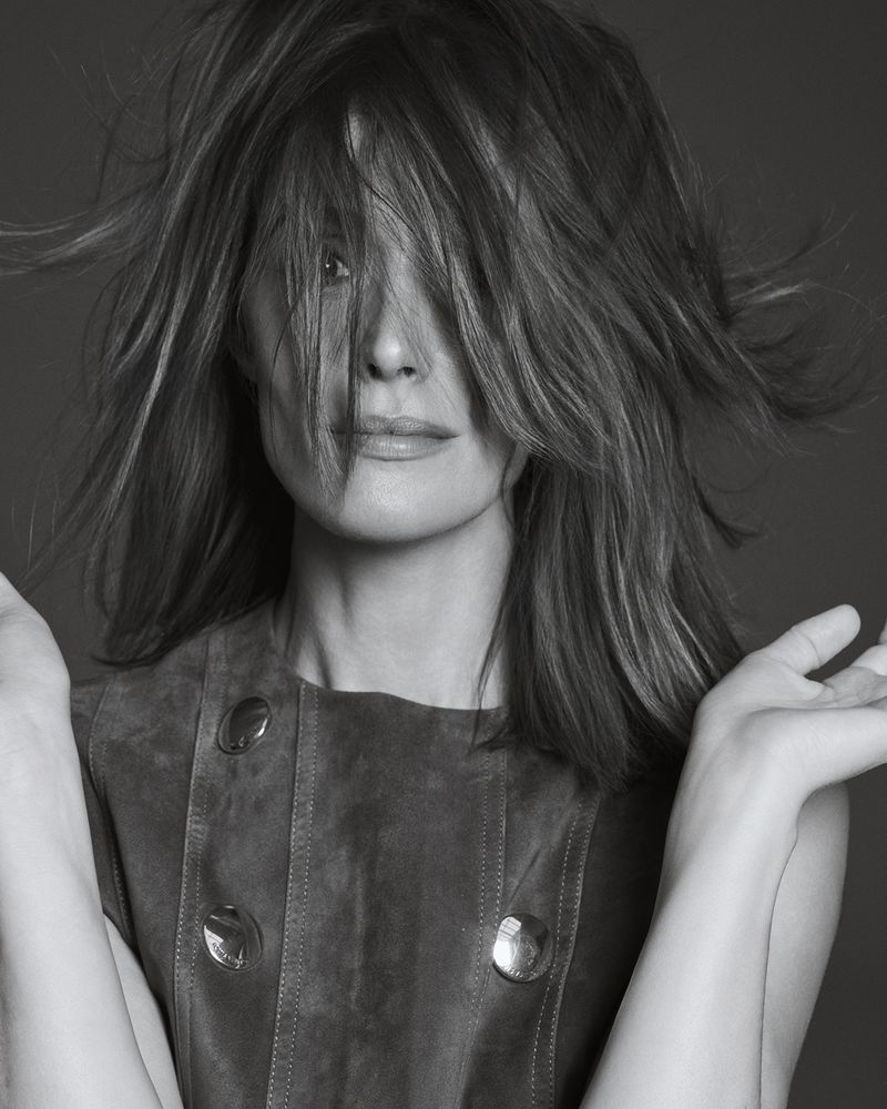 Black and white portrait of an adult female with medium-length hair covering her face, wearing a buttoned vest and with open hands.