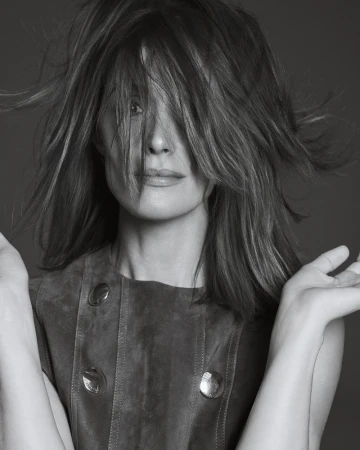 Black and white portrait of an adult female with medium-length hair covering her face, wearing a buttoned vest and with open hands.
