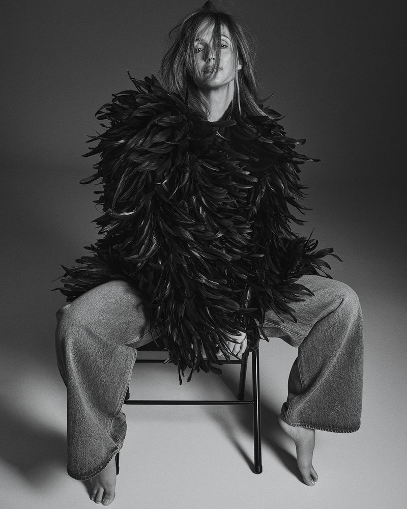 Black and white studio shot of a woman with feather boa, wide-legged jeans, and bare feet sitting on a simple stool, looking at camera.