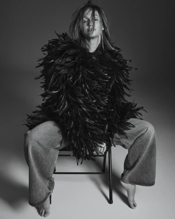 Black and white studio shot of a woman with feather boa, wide-legged jeans, and bare feet sitting on a simple stool, looking at camera.