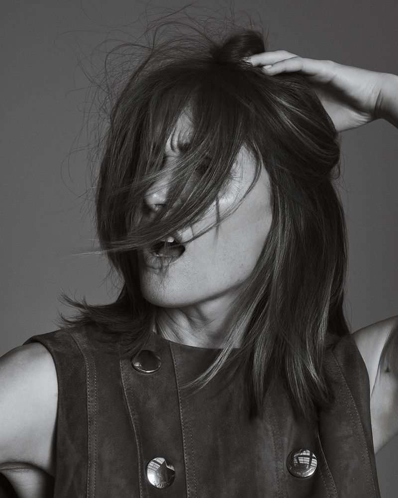 Monochrome portrait of an adult woman with tousled brown hair, open mouth, wearing a vest, and a neutral studio background.