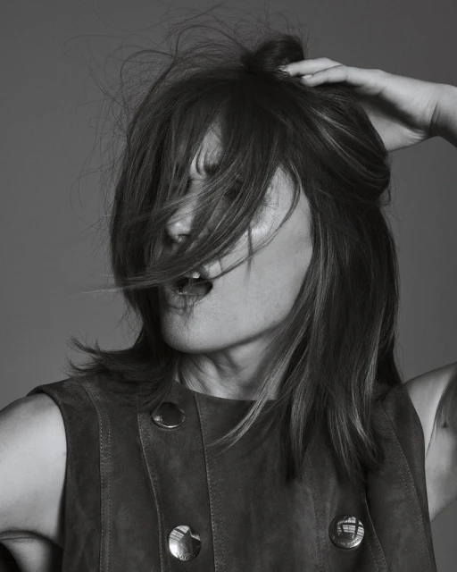 Monochrome portrait of an adult woman with tousled brown hair, open mouth, wearing a vest, and a neutral studio background.