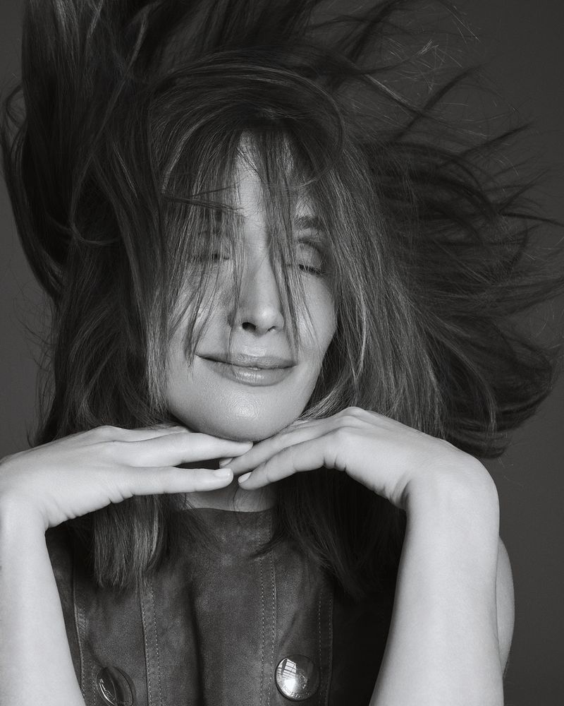 Black and white close-up portrait of Rose Byrne smiling with windblown hair and hands resting gently beneath her chin.