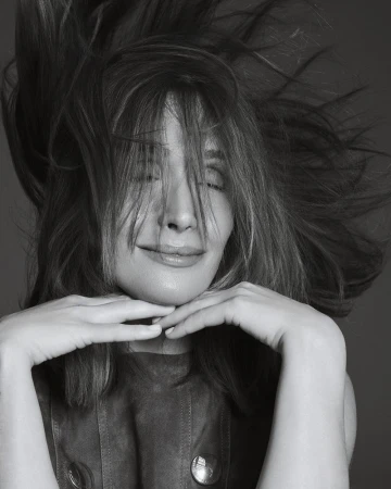 Black and white close-up portrait of Rose Byrne smiling with windblown hair and hands resting gently beneath her chin.