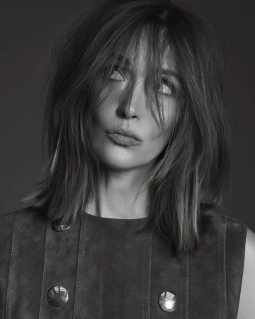 Monochrome portrait of a fair-skinned woman with shoulder-length hair, a sleeveless dress, and an expressive face, set against a plain backdrop.