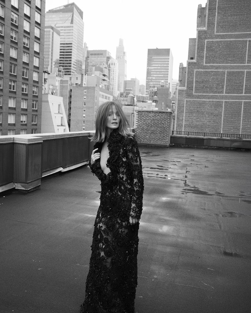 A woman in a black lace gown poses on a rooftop with a cityscape in the background, creating a striking black-and-white fashion image.