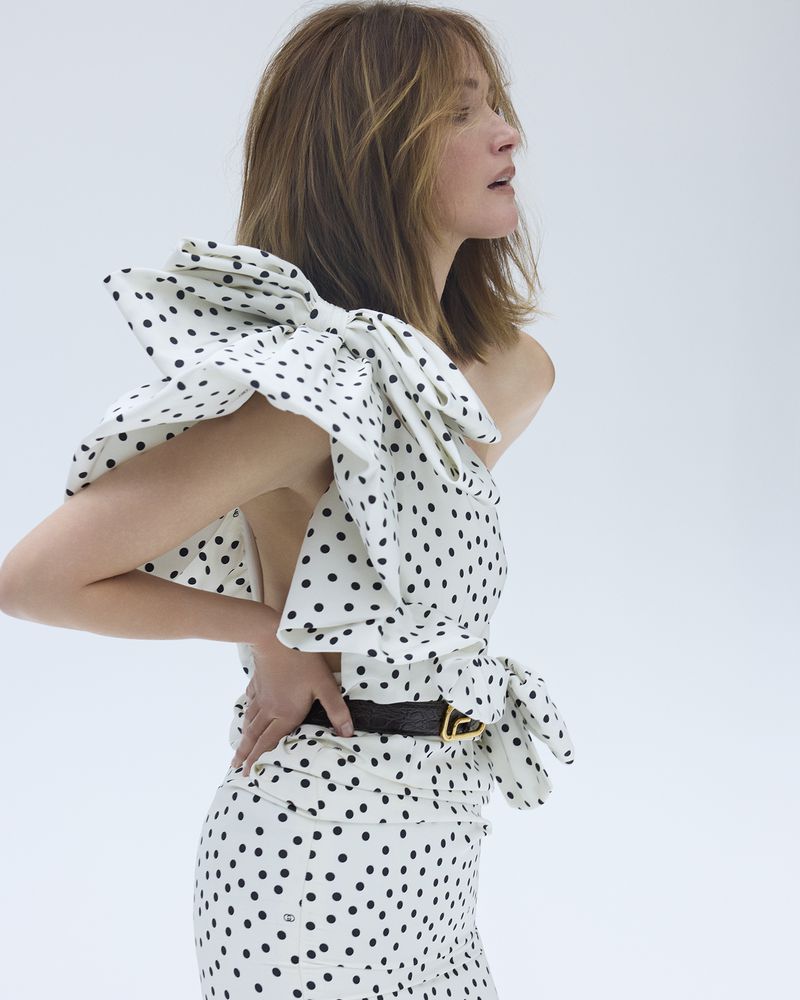Woman in polka dot dress with bow detail poses in a light studio. The dress has one shoulder and a form-fitting silhouette.