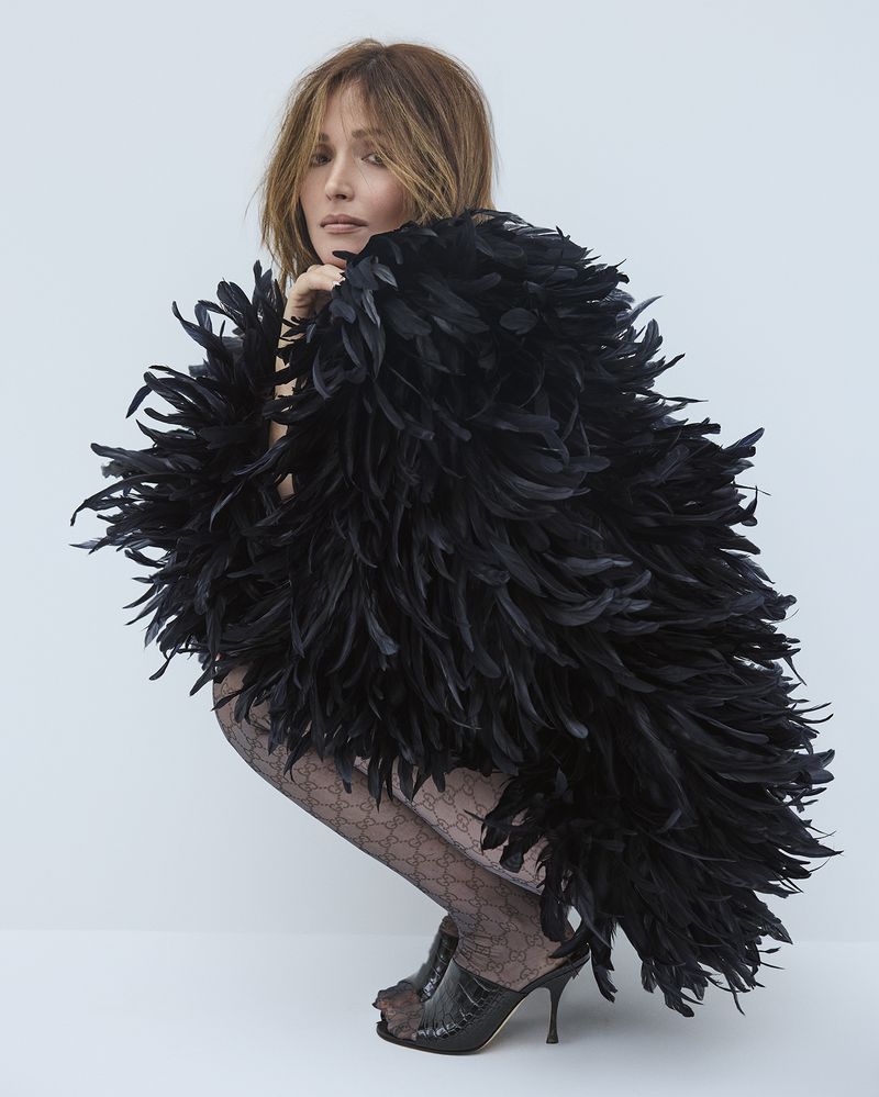 Adult female model in feathered black cape, patterned stockings, and heels posing in a studio with a neutral background.