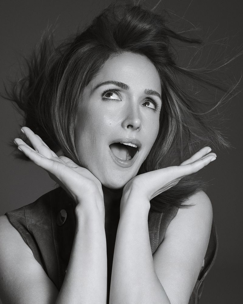 Portrait of a happy woman with wind-blown hair, hands framing face, looking upward against a neutral background in black and white.