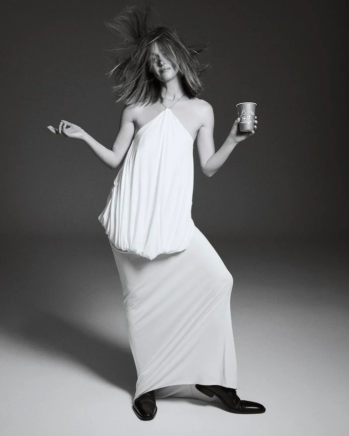 Rose Byrne in a white halter dress with hair flying, holding a cup against a grey studio backdrop